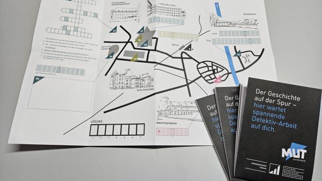 Auf einem Tisch liegt ein aufgeklapptes Entdeckungsheft. Darauf ist eine gezeichnete Karte und ein Kreuzworträtsel zu sehen. Am rechten Bildrand liegen drei weitere Exemplare des Entdeckungsheftes. Sie sind zugeklappt und tragen den Titel "Der Geschichte auf der Spur - hier wartet spannende Detektiv-Arbeit auf dich".. 