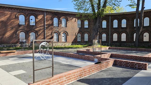 Der Gedenkort am Brückenkopf mit niedrigen Mauern, darauf Tafeln mit Illustrationen, im Hintergrund das ehemalige Hafthaus
