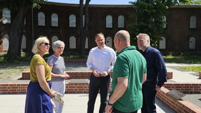 Gruppe von fünf Besuchenden stehen am neuen Gedenkort am Brückenkopf und sind in ein Gespräch vertieft.