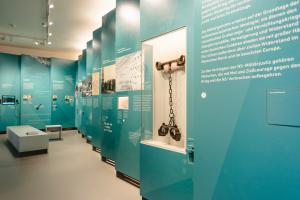 Das Foto zeigt einen Blick in die Ausstellung "Mut und Ohnmacht": Die Wände sind mit türkis-blauen Tafeln gestaltet, auf denen Texte und großformative Bilder abgedruckt sind. Im Vordergrund hängt eine Hand- und Fußfessel aus Metall.