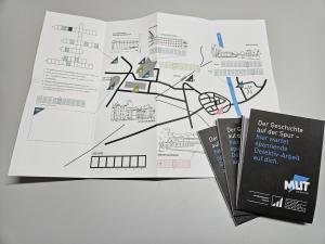 Auf einem Tisch liegt ein aufgeklapptes Entdeckungsheft. Darauf ist eine gezeichnete Karte und ein Kreuzworträtsel zu sehen. Am rechten Bildrand liegen drei weitere Exemplare des Entdeckungsheftes. Sie sind zugeklappt und tragen den Titel "Der Geschichte auf der Spur - hier wartet spannende Detektiv-Arbeit auf dich".. 